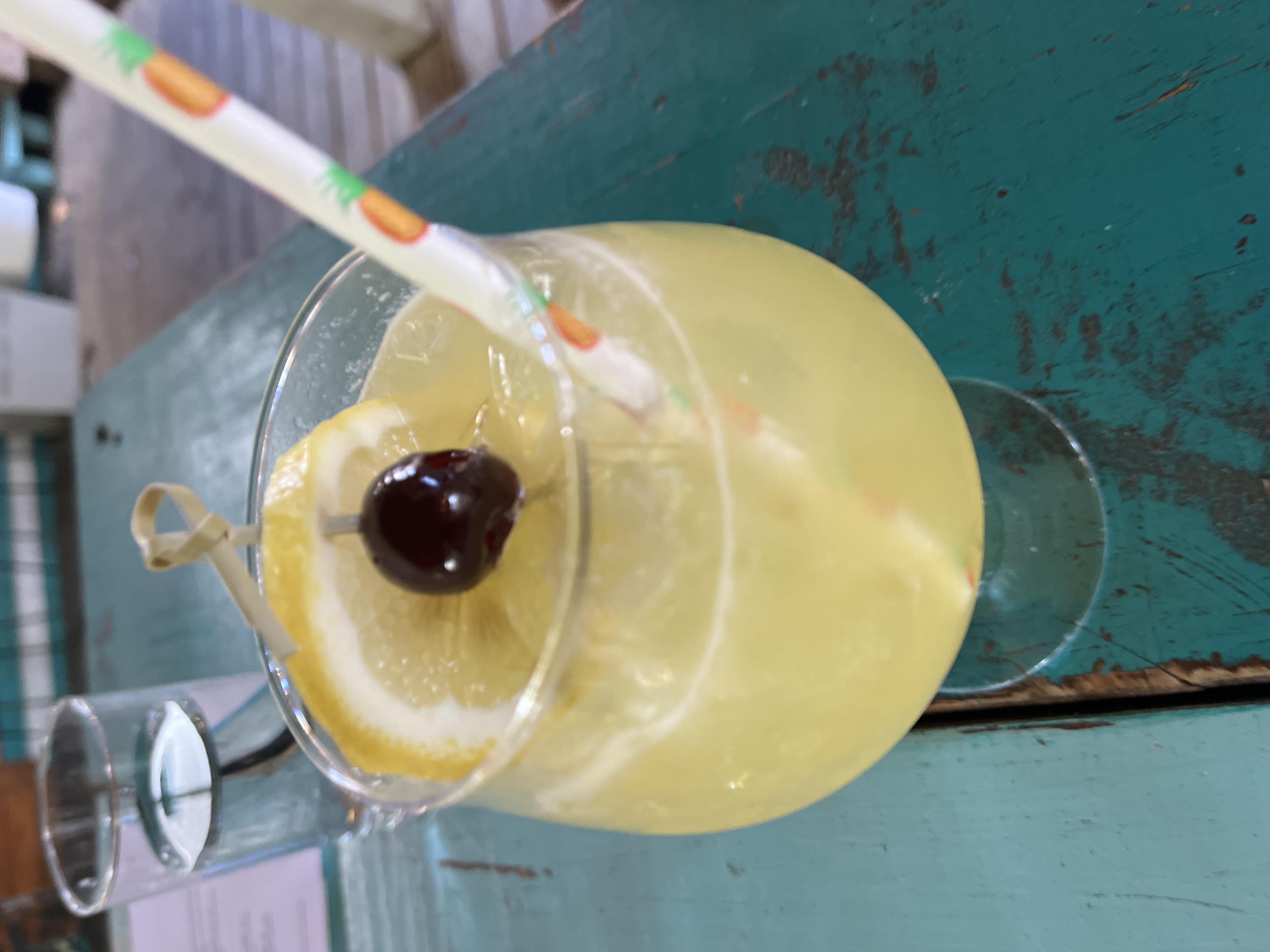 Yellow cocktail with lemon slice, cherry, and striped straw on a turquoise table.