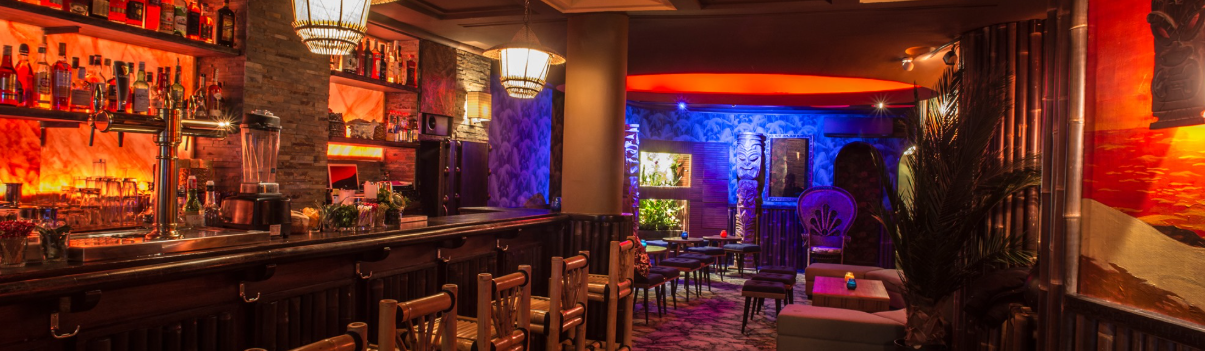 Cozy bar with colorful lighting, wooden chairs, and shelves filled with bottles.