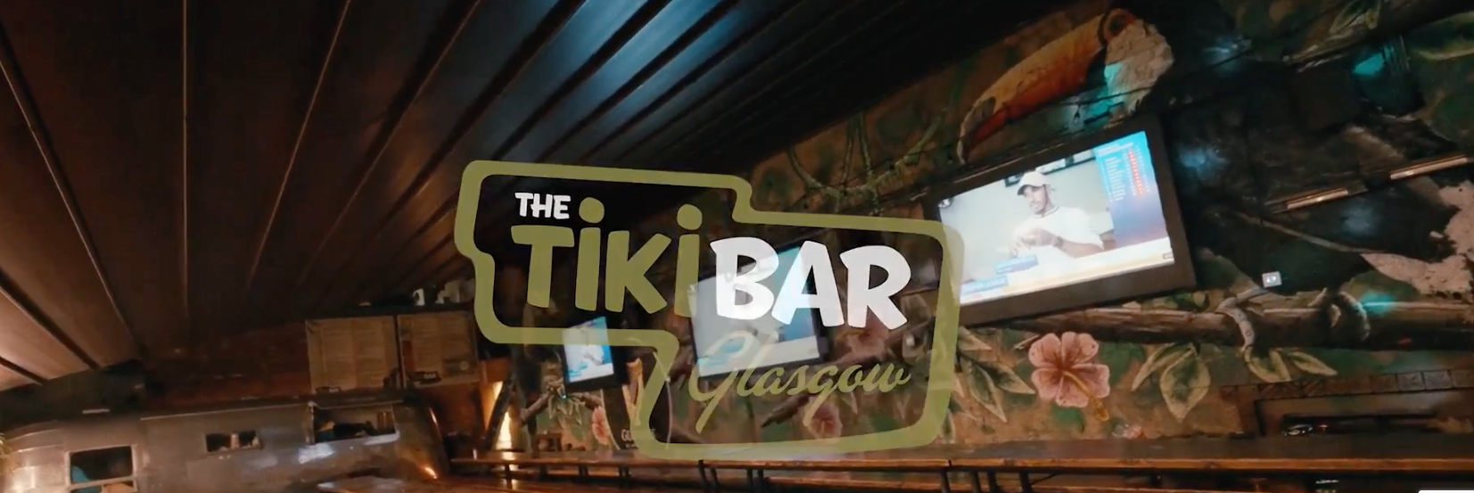 Tiki bar interior with tropical decor and wall-mounted screens.