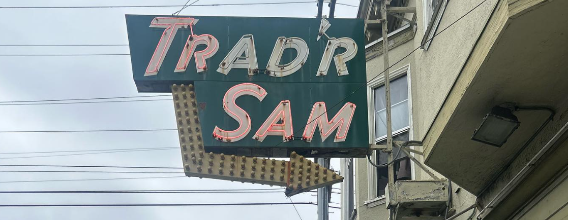 A vintage sign reads "TRADR SAM" with an arrow, against a cloudy sky.