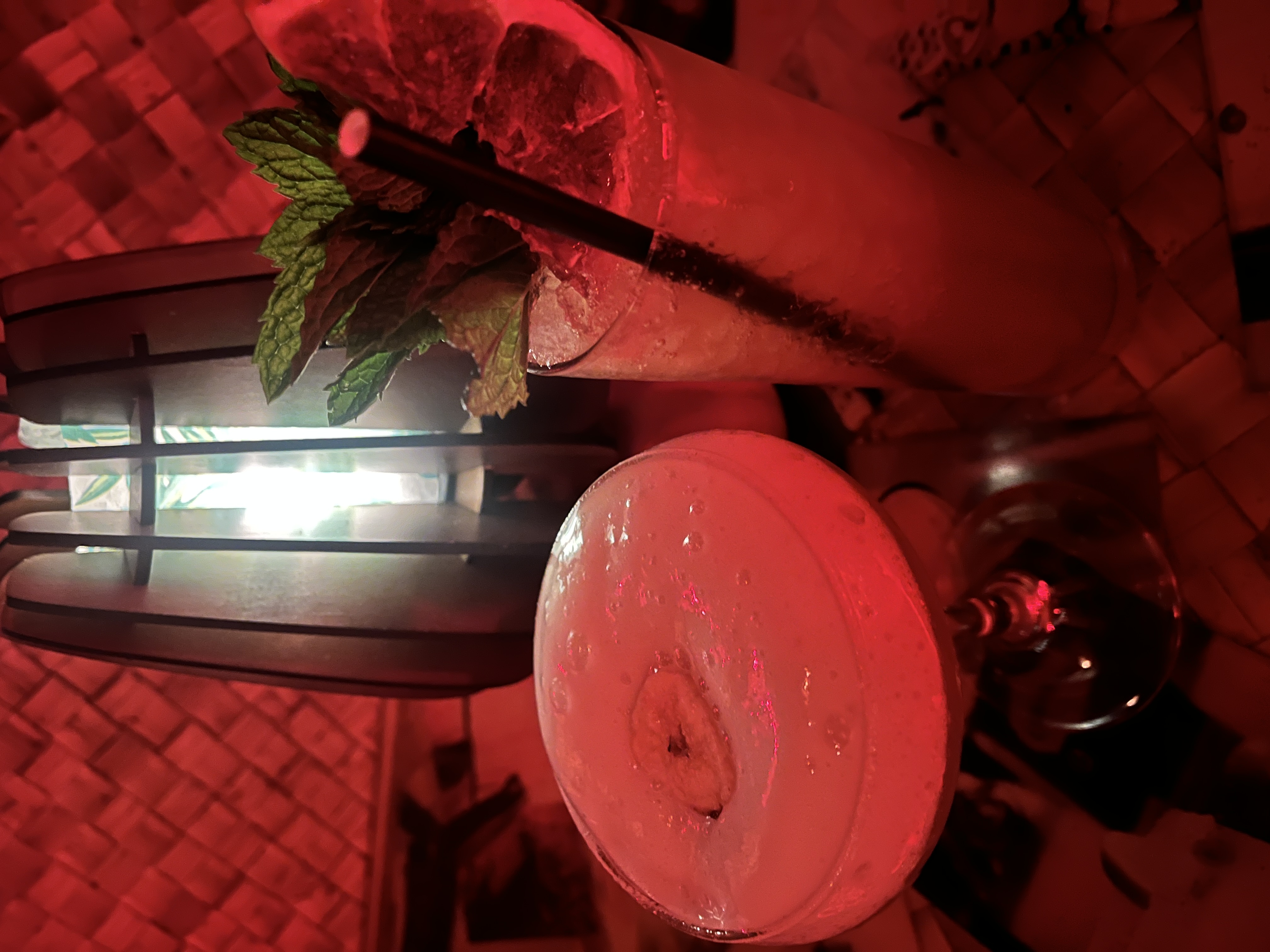 Two cocktails on a table with red lighting, garnished with mint and fruit.