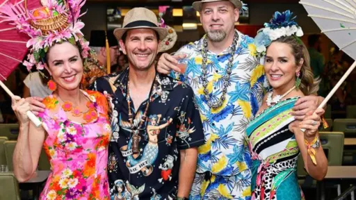 Two men and two women in colorful Hawaiian attire, smiling, holding umbrellas indoors.