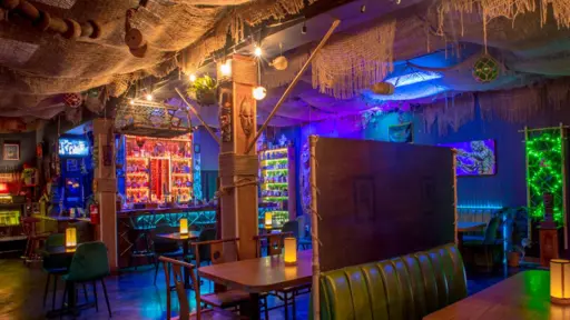 Dimly lit, colorful bar with booths and neon lights, creating a cozy, eclectic atmosphere.